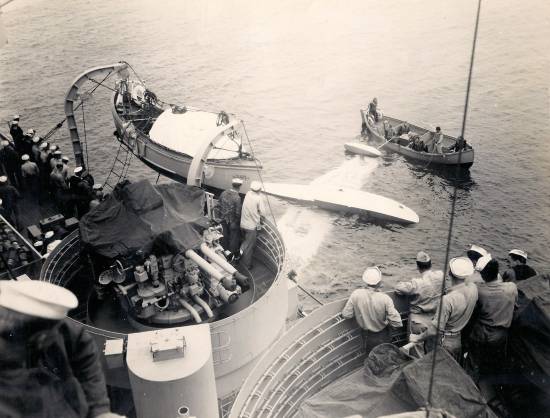 We see a wing lying flat in the water. Two wing floats are visible, but they thrust up. An OS-2U Kingfisher may be upside down, as members of the crew of the USS Pasadena look on, and their whaleboat