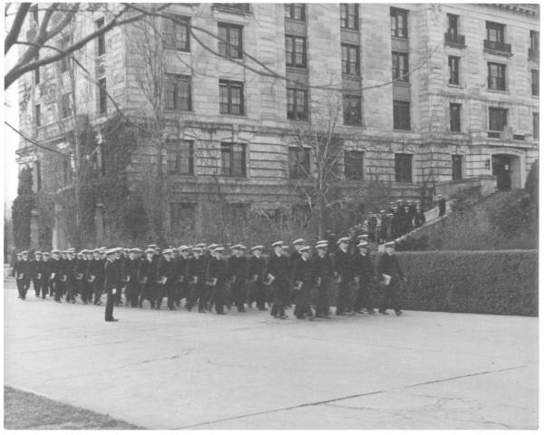 About 1938, at the U.S. Naval Academy, Midshipman in service blue uniforms carrying book(s), marching behind a wing of Bancroft Hall.